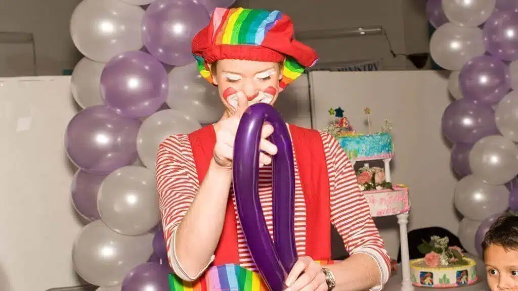 Balloon twister in a clown costume twisting a long purple balloon at a birthday party