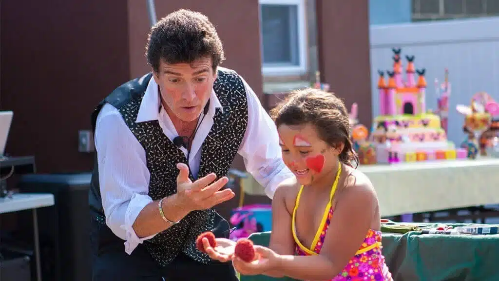 Kids magician doing a trick with a young girl at an outdoor birthday party