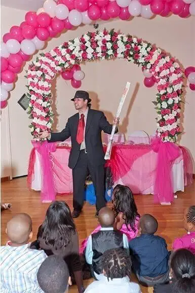 Kids magician performing on stage under a balloon arch at a party hall
