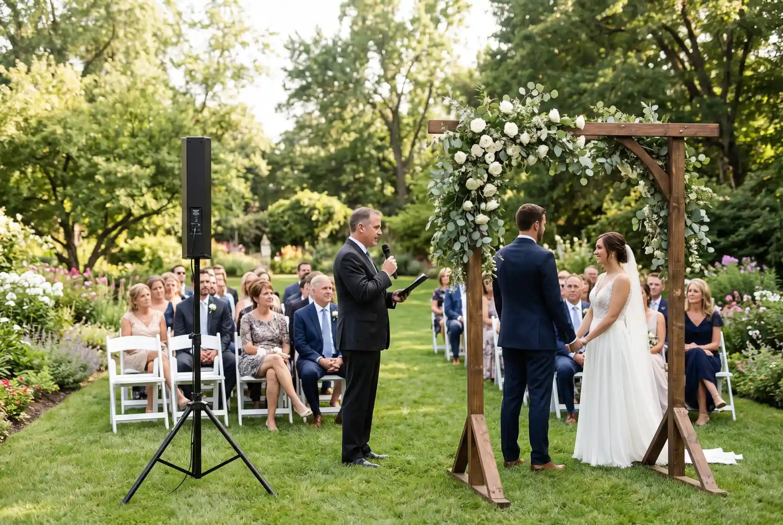 Officiant speaking into a wireless microphone at an outdoor garden wedding ceremony with a speaker on a stand beside the aisle