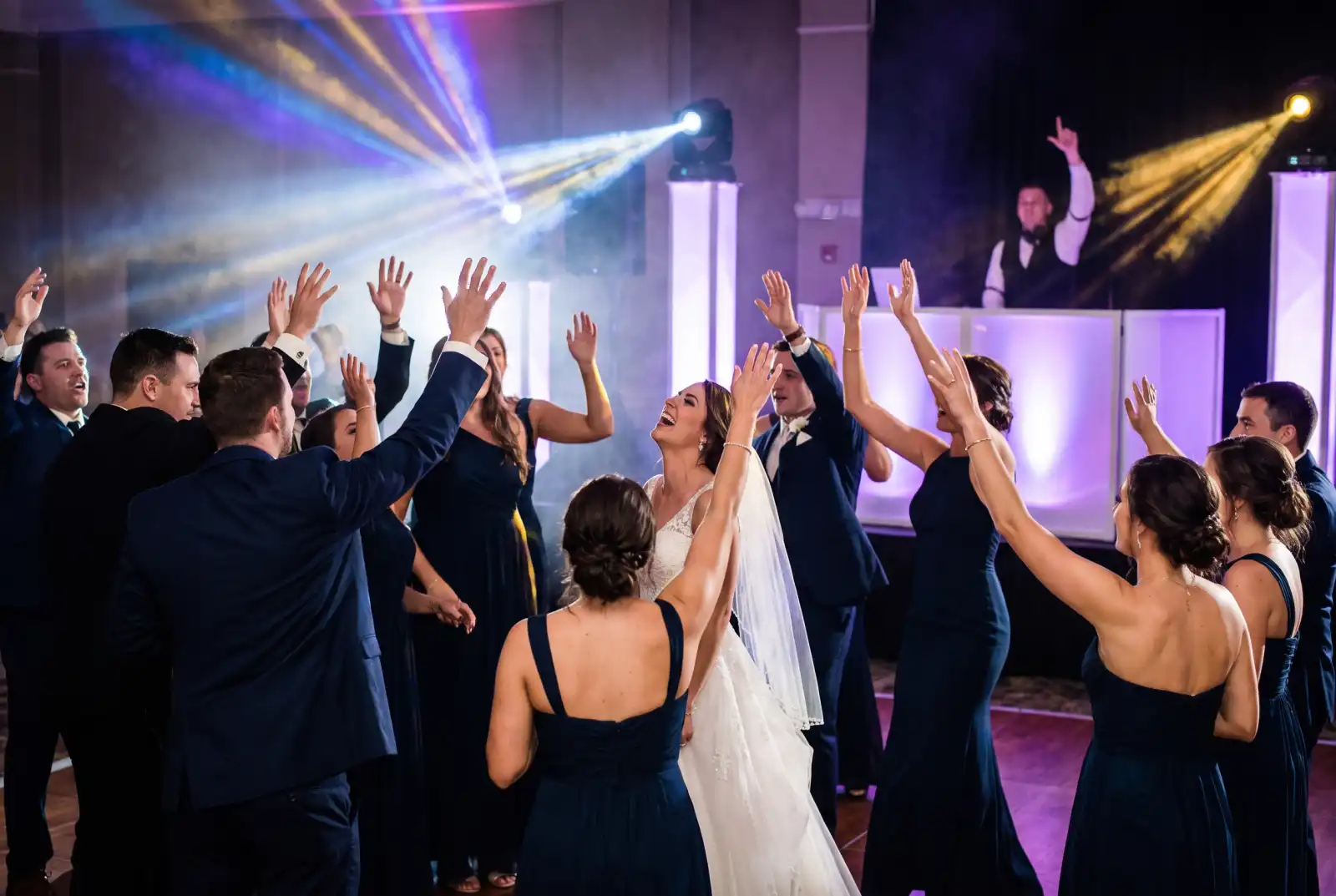 Wedding guests jumping with hands up on a packed dance floor, moving head beams and haze, DJ raising his hand at a white DJ booth