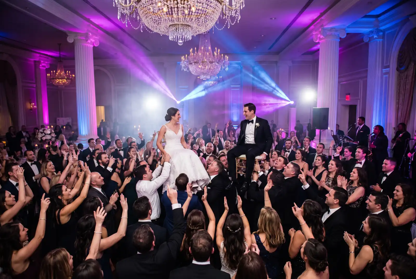 Bride and groom lifted on chairs during the hora while guests dance in a circle under purple uplighting and moving head beams