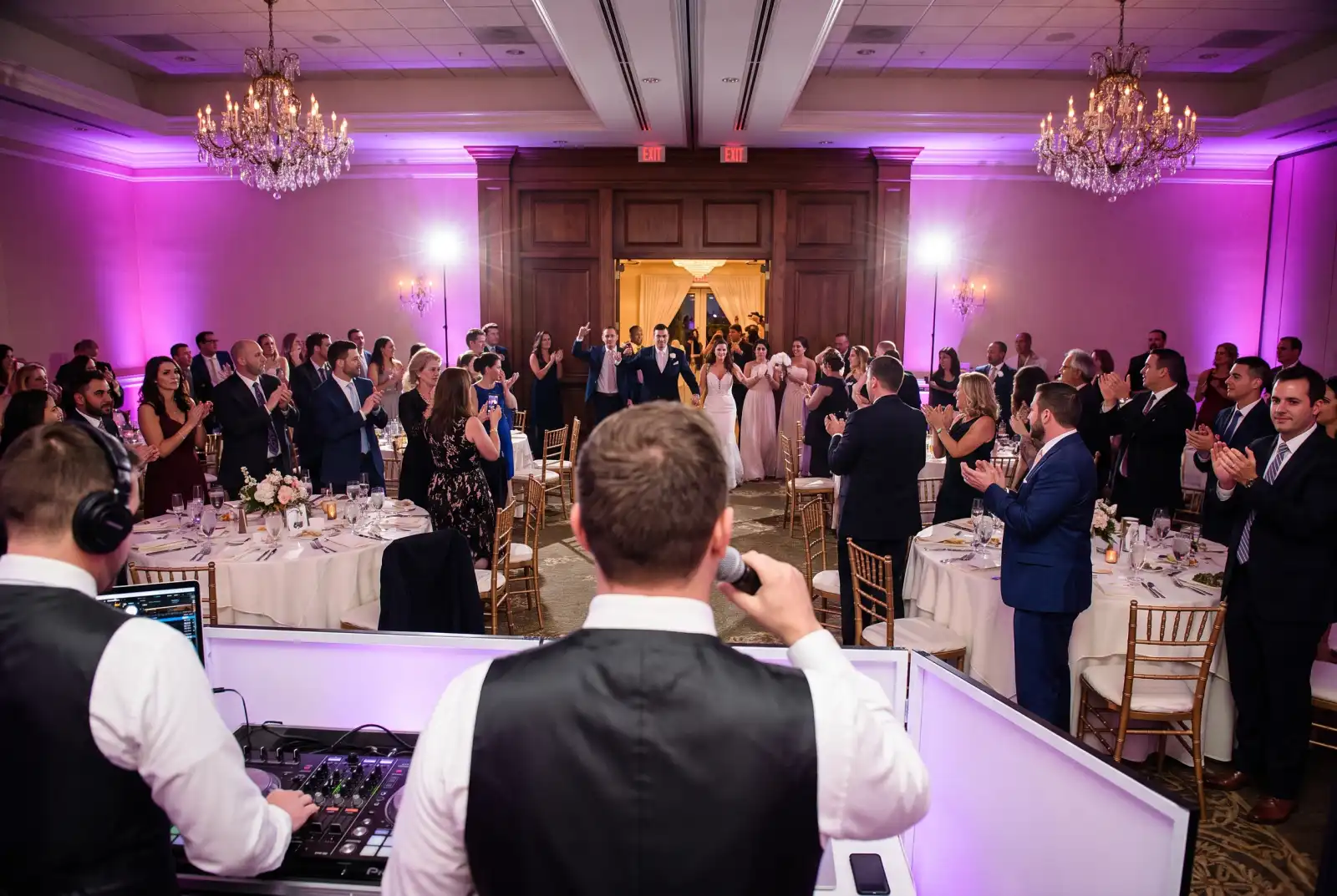 DJ and MC with a wireless microphone at a white DJ booth announcing the bridal party entrance as guests stand and clap in a ballroom with purple uplighting