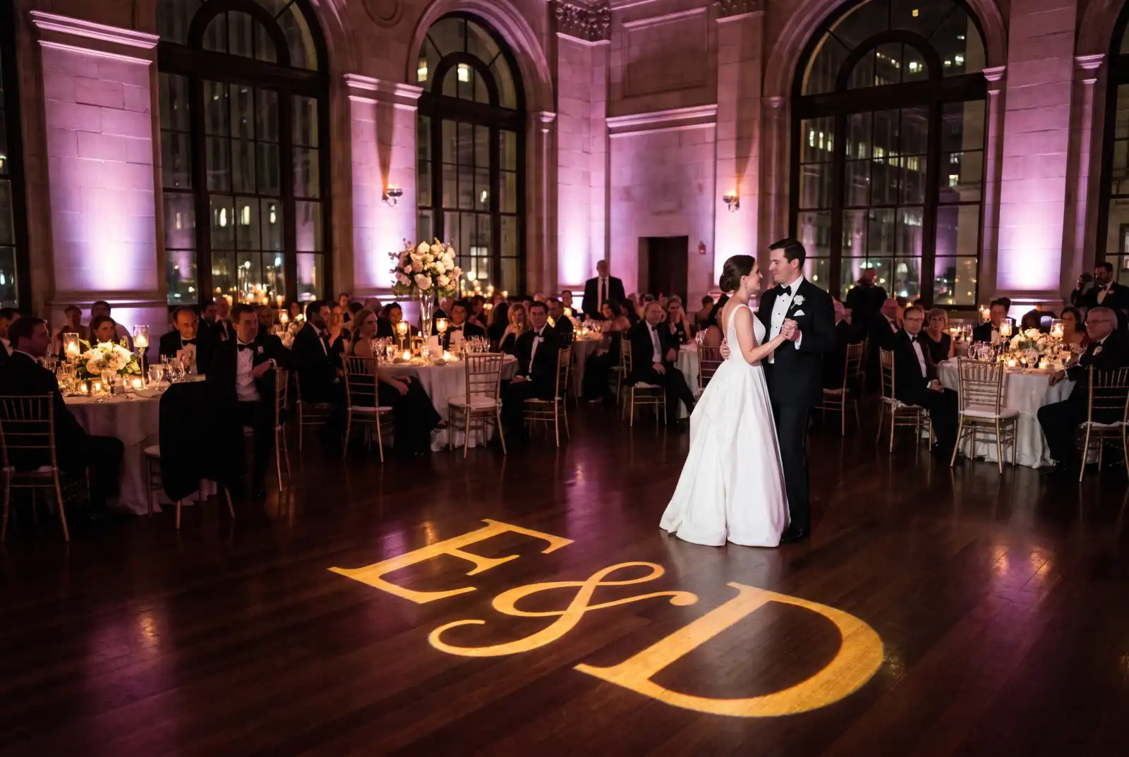 Gold monogram with the couple initials projected on a wooden dance floor at a wedding reception with purple uplighting