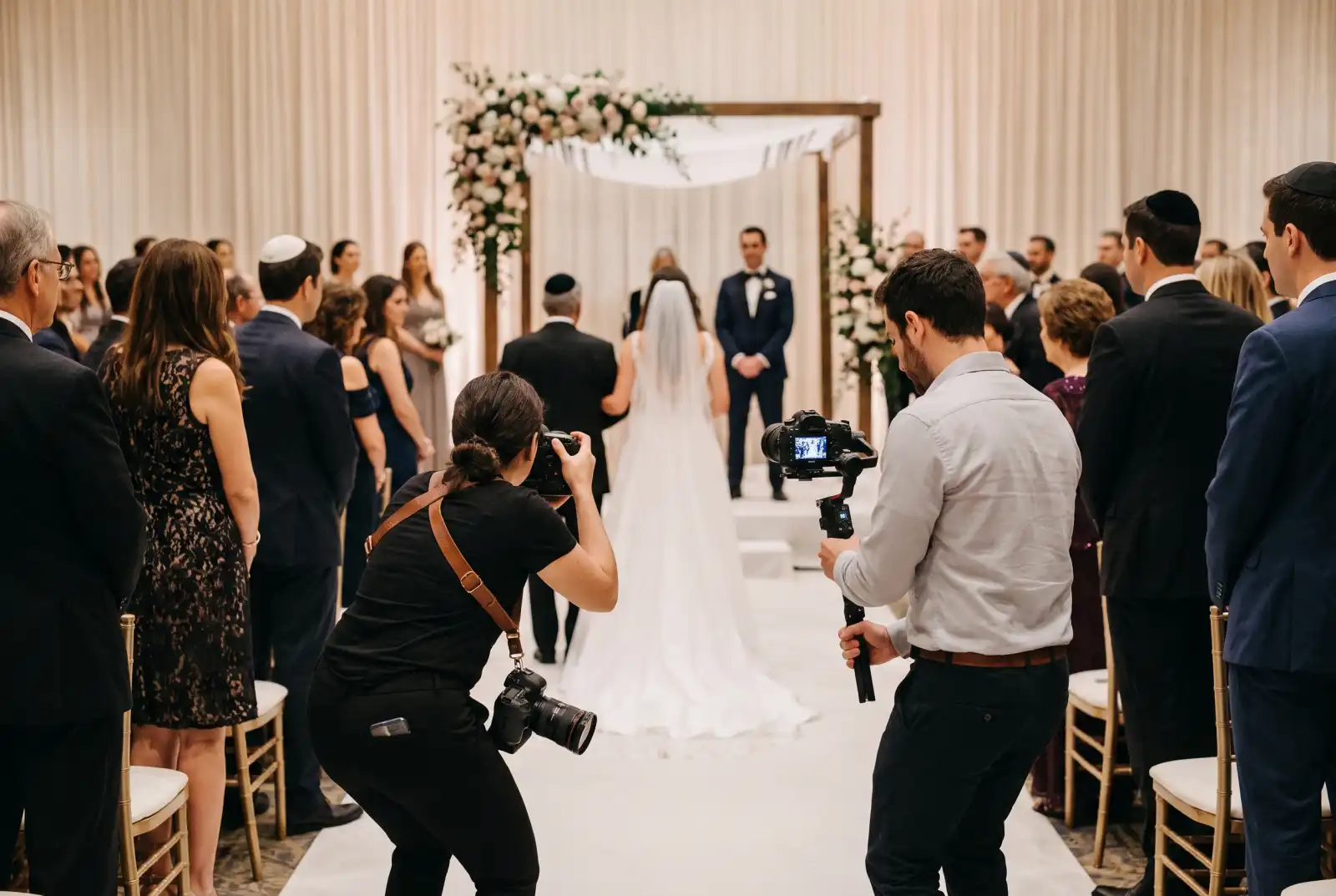 Wedding photographer and videographer with a gimbal filming the bride walking down the aisle to the chuppah where the groom waits