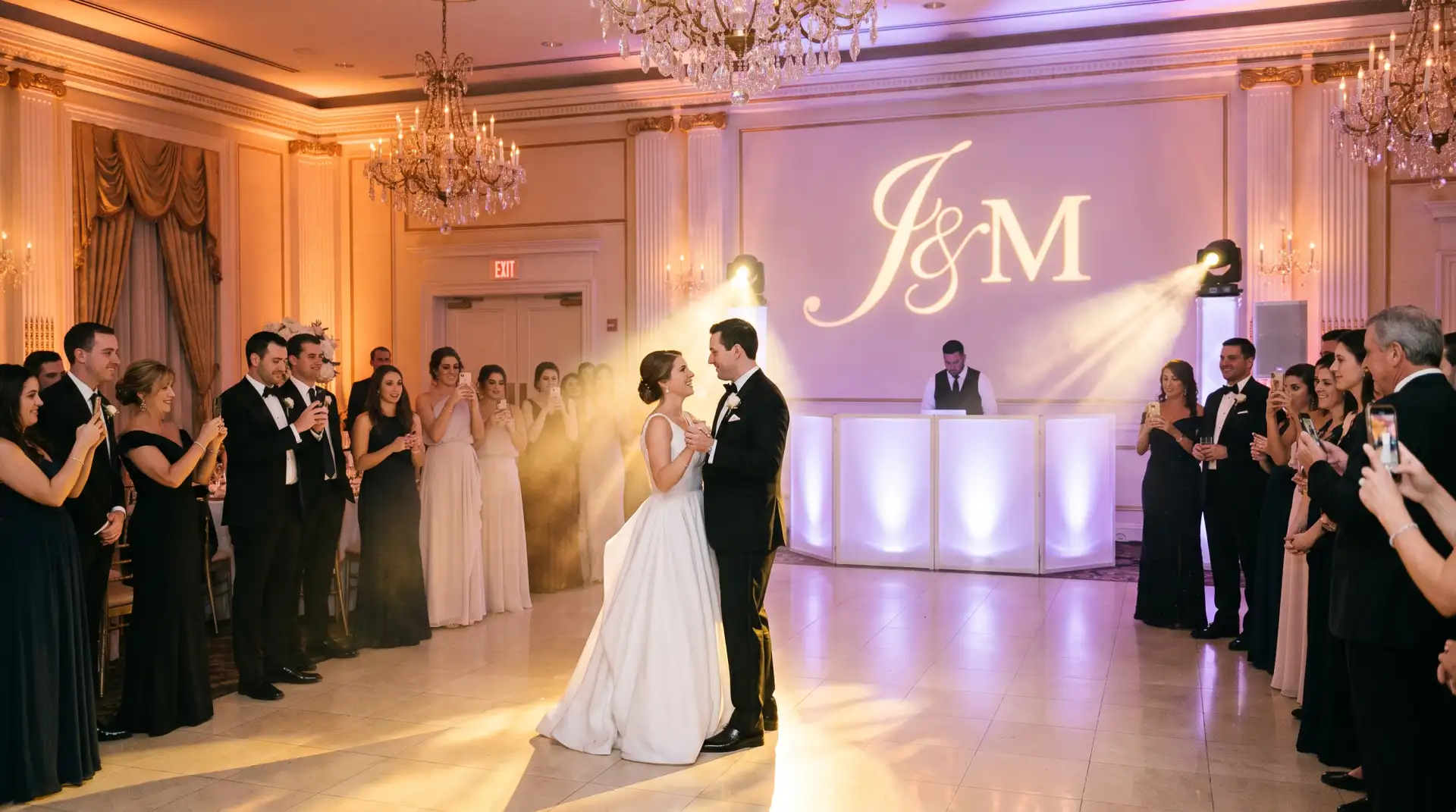 Bride and groom share their first dance in a NYC ballroom with amber and purple uplighting, a gold monogram light, chandeliers and a lit white DJ booth