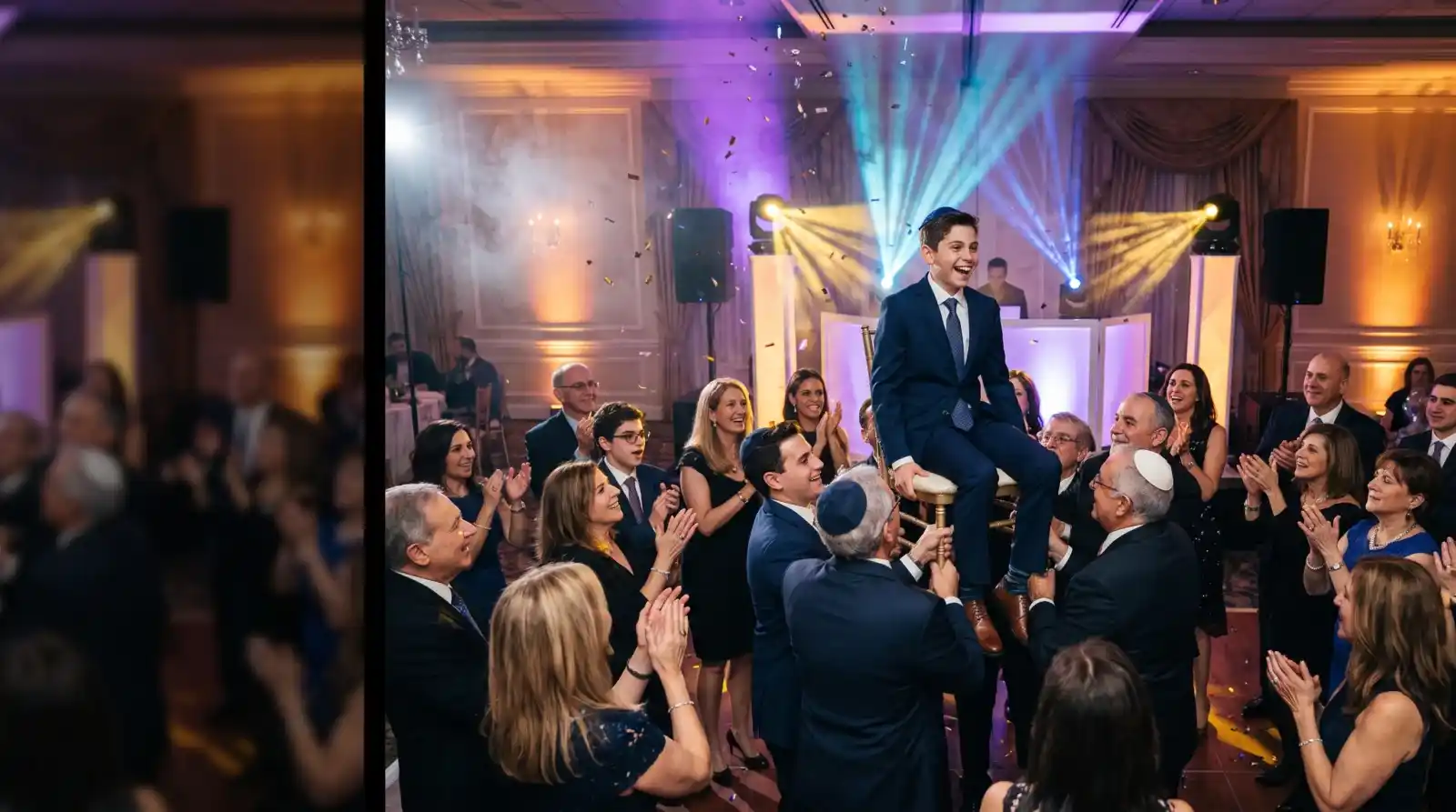 Bar mitzvah boy lifted on a chair during the hora, surrounded by dancing family under purple and gold moving-head lighting with a DJ booth behind