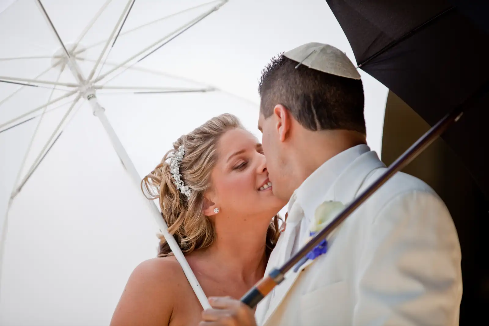 Bride and groom in a white suit and kippah sharing a kiss under a white umbrella in a wedding portrait