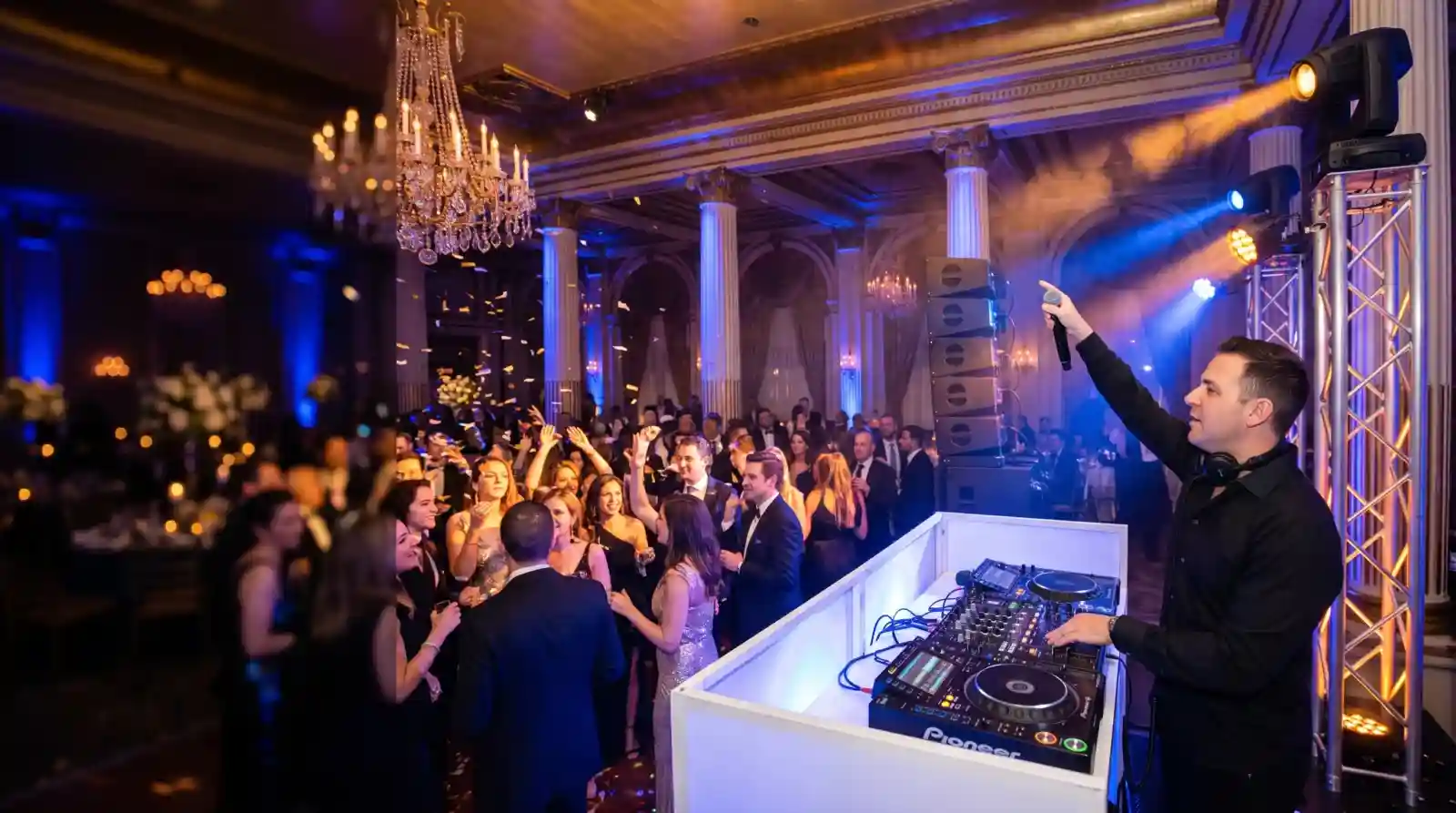DJ NYC: DJ at a white DJ booth raising a wireless microphone over a packed ballroom dance floor, with moving head lights on truss and chandeliers