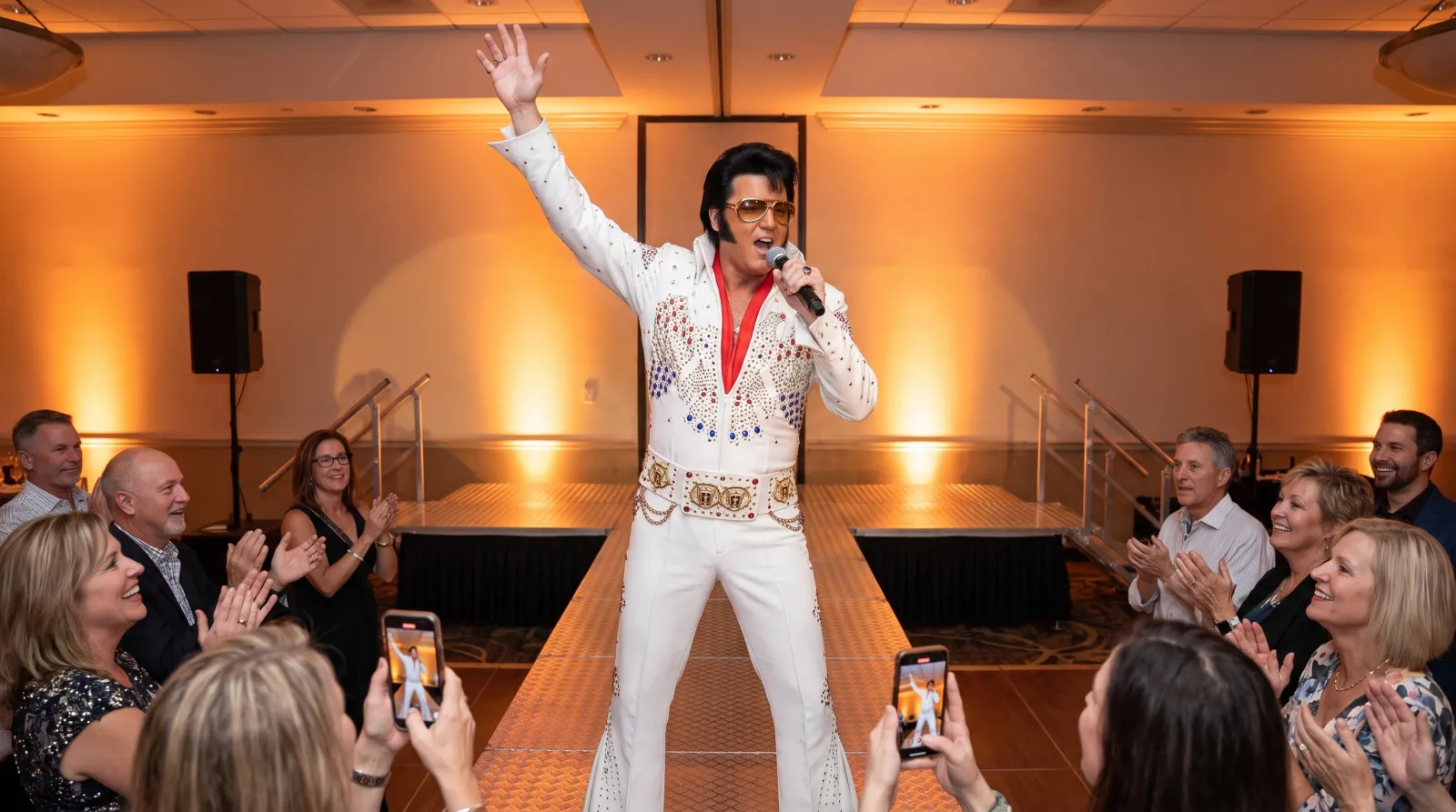 Elvis impersonator singing on an aluminum stage at a party while guests clap and film