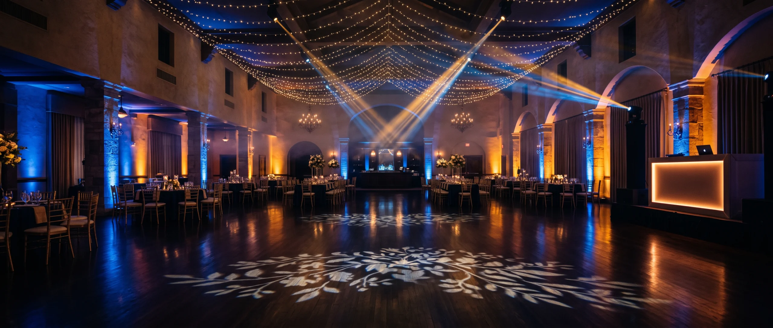 Wedding Rentals NYC: centered view of a ballroom lit with blue and amber uplighting, ceiling string lights, light beams and a leaf gobo on the polished dance floor