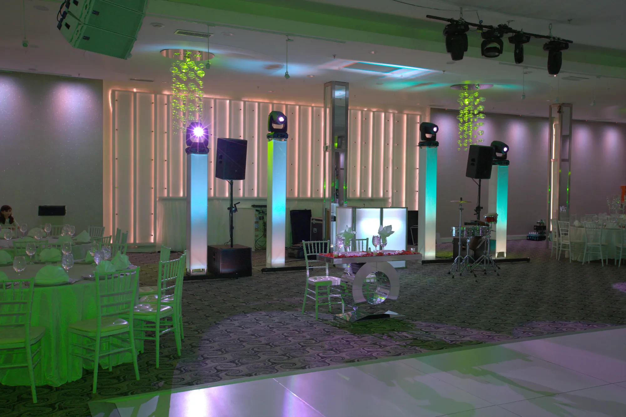 Ballroom with a lit DJ booth, two powered speakers on lit pedestals, a subwoofer and moving head lights