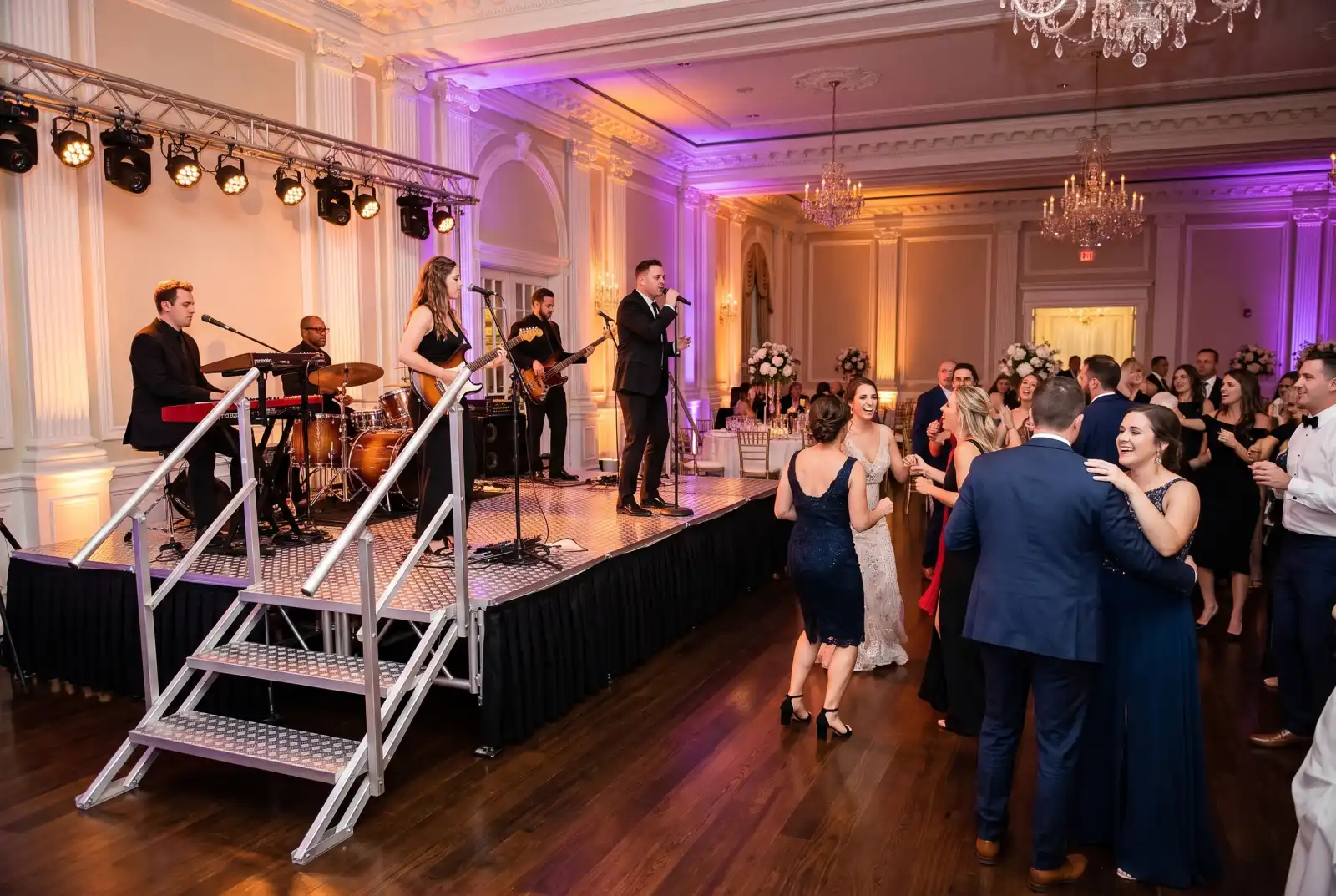 Live band on an aluminum portable stage with black skirting and handrail steps as guests dance at a wedding reception