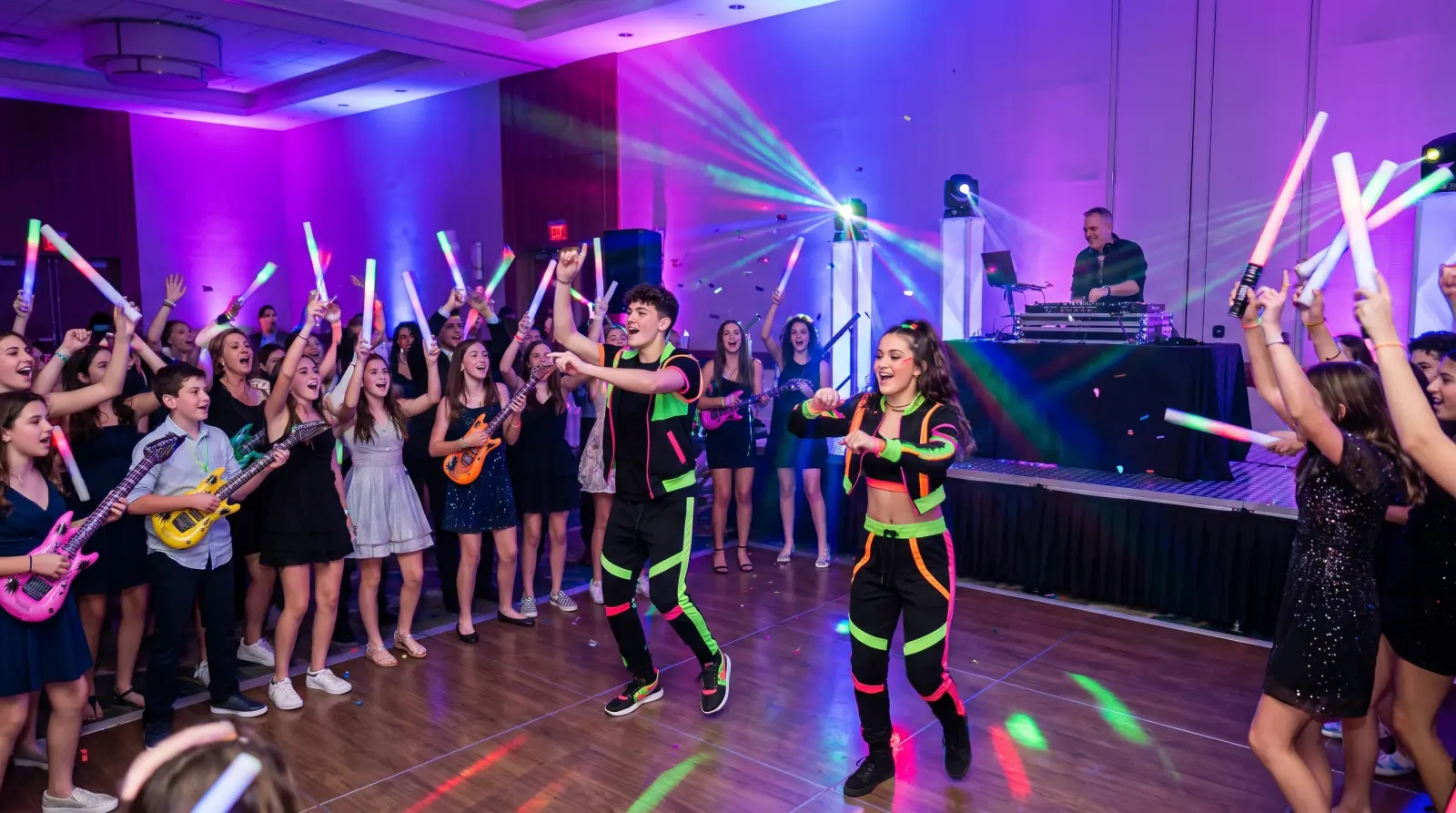Party dancers leading teens with glow sticks on a bat mitzvah dance floor, with the DJ behind them