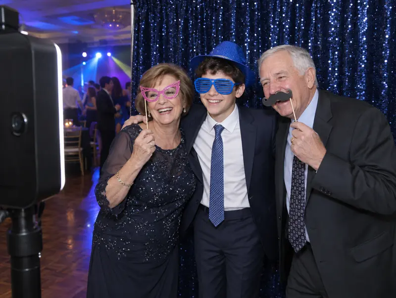Photo booth at a bar mitzvah: boy and grandparents posing with props in front of a navy sequin backdrop