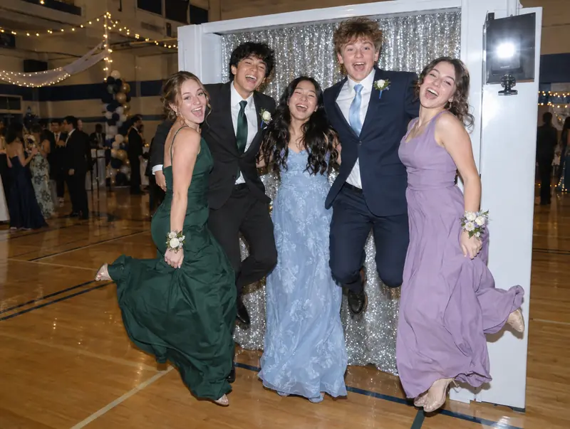 Photo booth at a high school prom: teens jumping in front of a silver backdrop in a decorated gym