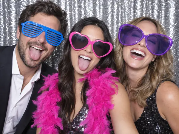 Photo booth print: three friends in oversized party sunglasses and a pink feather boa making silly faces