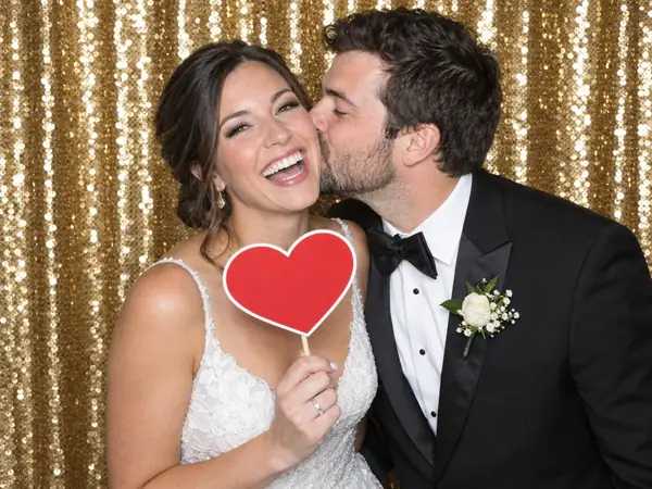 Photo booth rental print: bride holding a heart prop while the groom kisses her cheek in front of a gold sequin backdrop