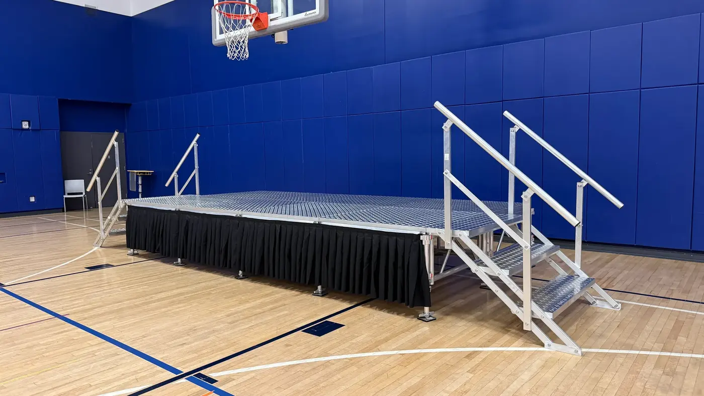 Side view of a portable stage with black skirting and handrail steps on a gymnasium wood floor