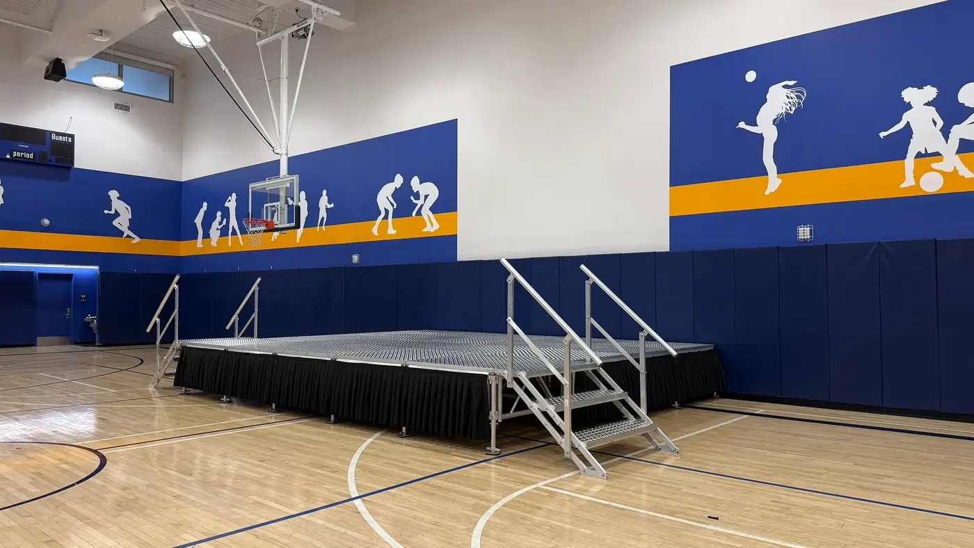 School Event Rentals: angled view of a portable stage with black skirting and handrail steps on a gymnasium sports floor