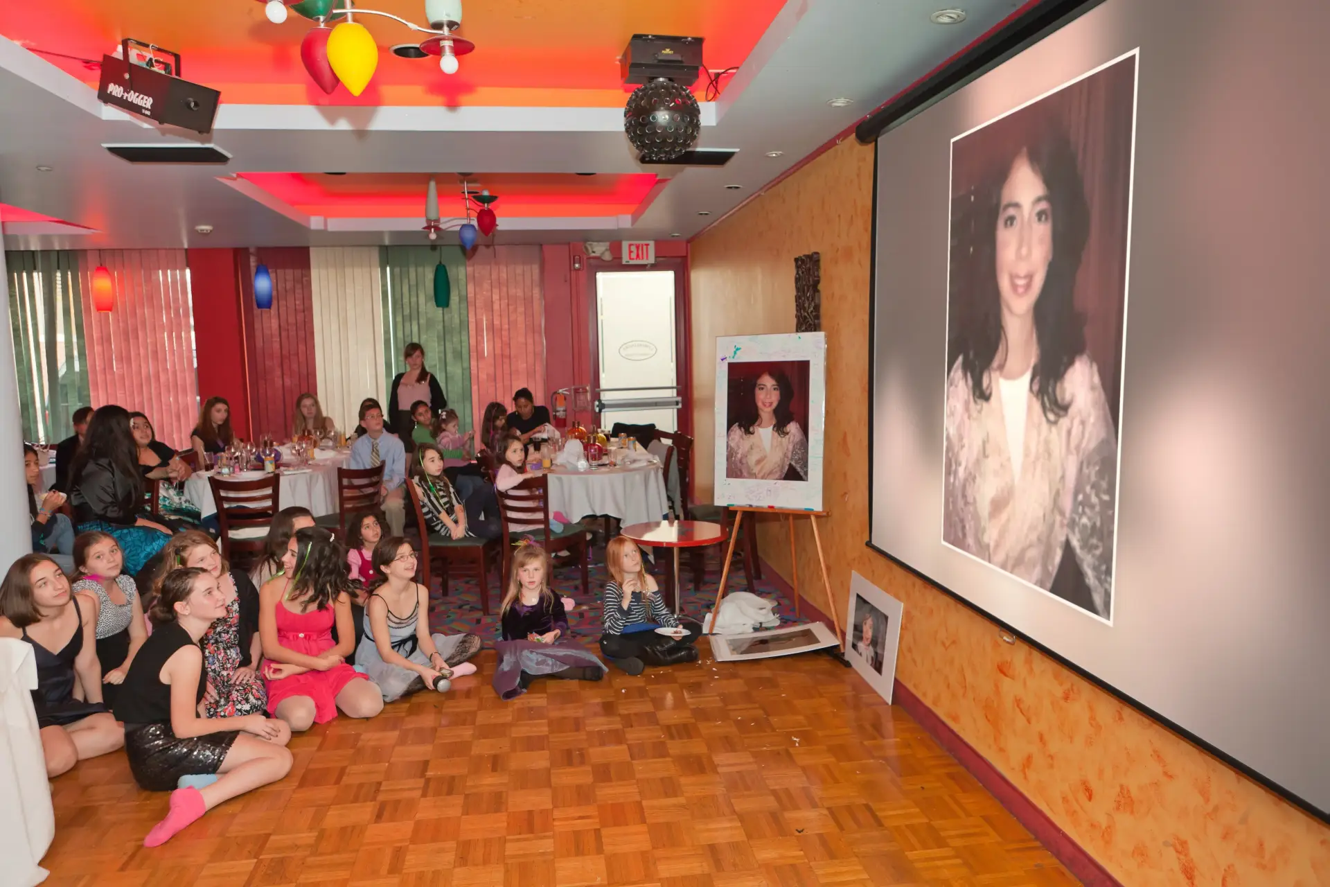 Girls sitting on the floor watching a photo montage on a large projection screen at a bat mitzvah