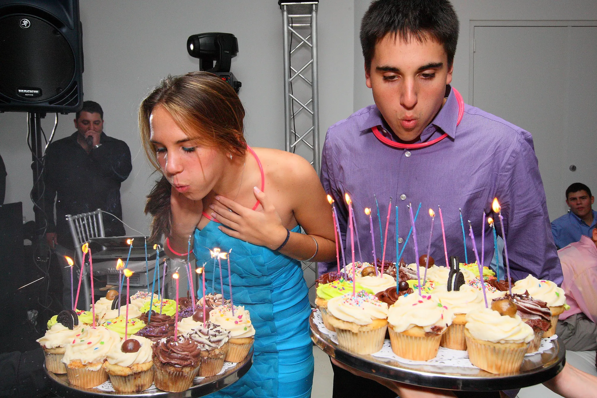 Teens blowing out candles on trays of cupcakes at a Sweet 16, with a moving head light on a truss behind them