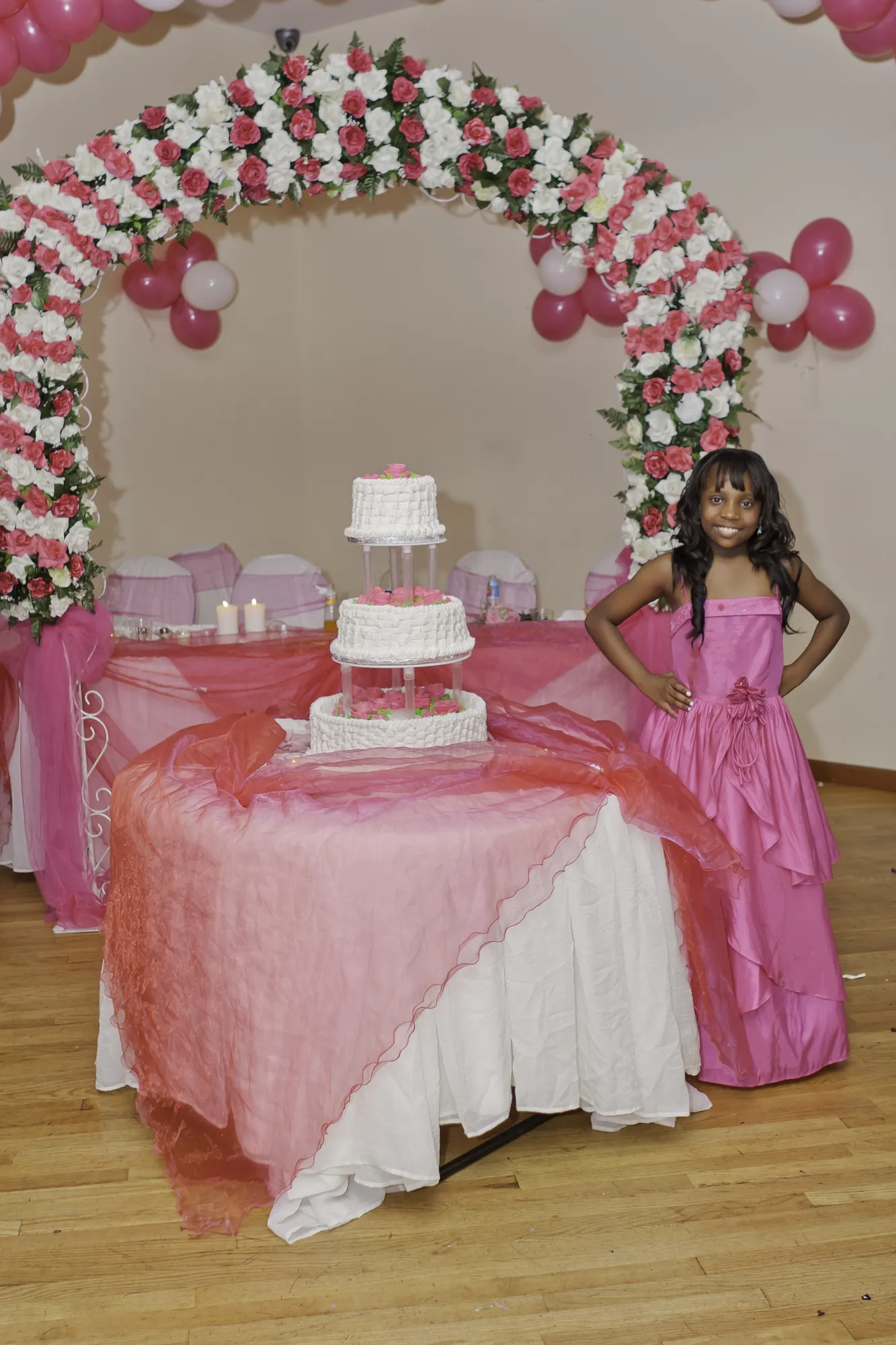 Sweet 16 girl in a pink gown beside her three-tier cake under a pink and white flower and balloon arch