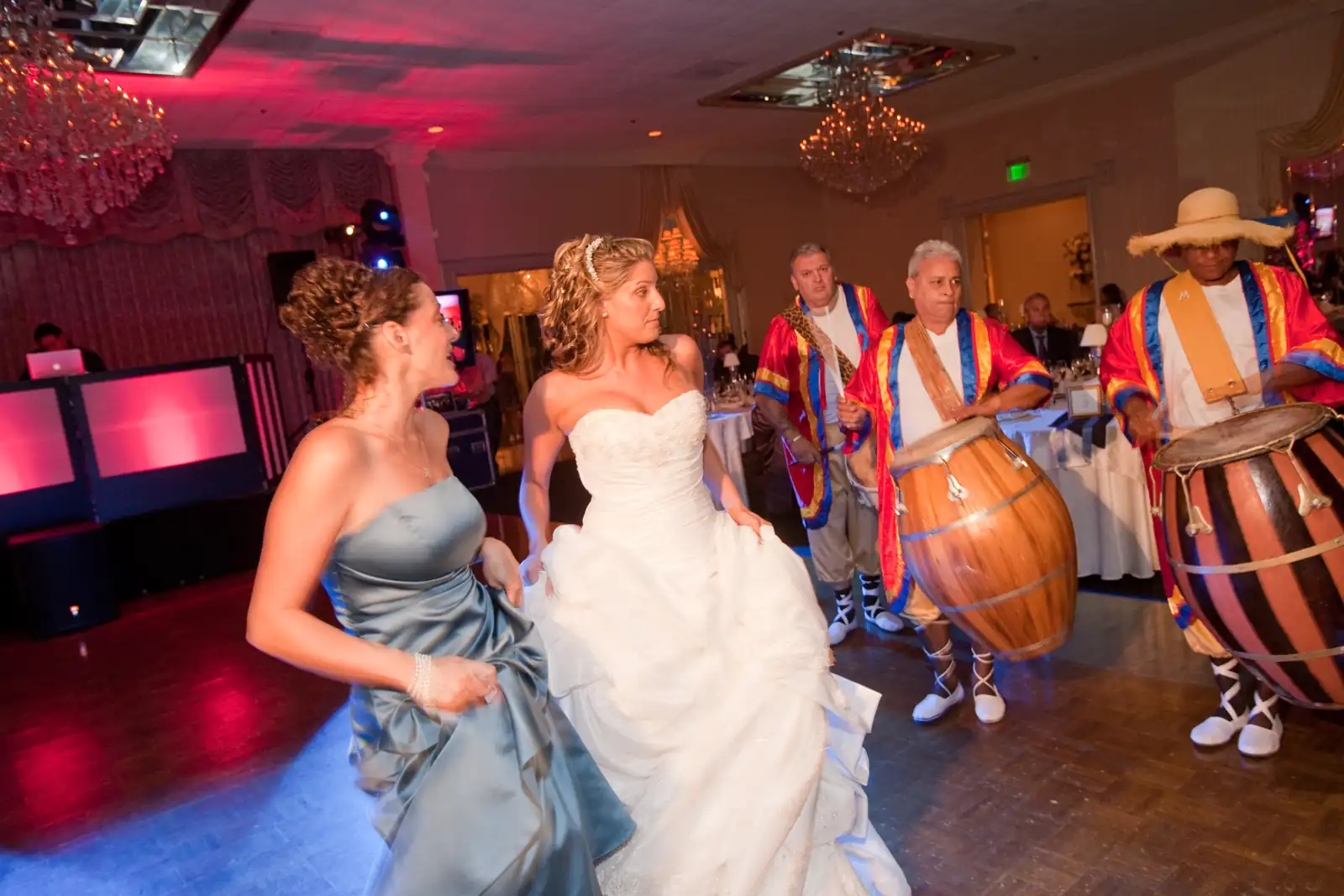 Bride and a bridesmaid dancing as costumed drummers play beside a lit DJ booth under red and pink uplighting